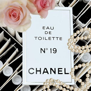 May include: A white card with the text "EAU DE TOILETTE N° 19 CHANEL" is surrounded by a pearl necklace and pink roses. The card is framed in silver. Small perfume vials are visible in the background.