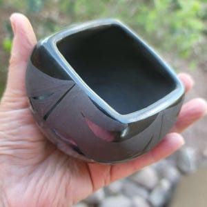Puede incluir: Un pequeño cuenco cuadrado de cerámica negra con acabado brillante. El cuenco presenta un diseño decorativo grabado en el exterior. El cuenco se sostiene en una mano, mostrando su tamaño y forma. El cuenco probablemente se utiliza con fines decorativos o funcionales.