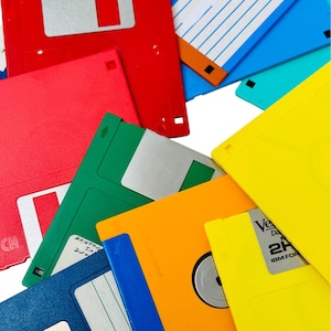 Puede incluir: Una colección de disquetes de colores, incluyendo rojo, azul, verde, amarillo y naranja. Los disquetes están dispuestos en un patrón aleatorio, con algunos superpuestos. Los disquetes tienen todos 8,9 cm de diámetro.