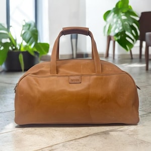 May include: A brown leather duffel bag with a top handle and a rectangular label. The bag has a rounded shape and a zipper closure. The bag is made of a smooth, textured leather.