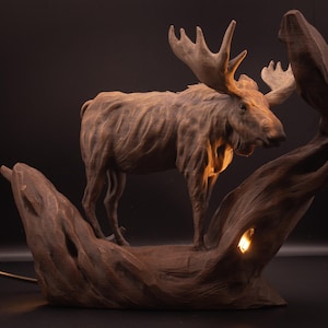May include: A detailed wooden sculpture of a moose with large antlers, standing on a carved wooden base. The sculpture has a warm brown colour and is illuminated by a soft light, creating a rustic and natural aesthetic. The lamp is approximately 46 cm tall.