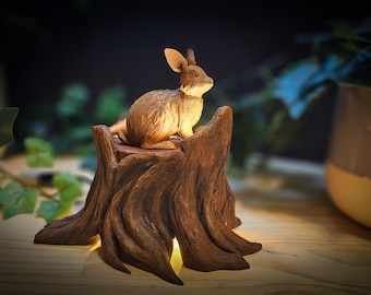 May include: A wooden rabbit figurine sits atop a carved wooden tree stump, illuminated from within. The stump has a rustic brown finish and intricate details.