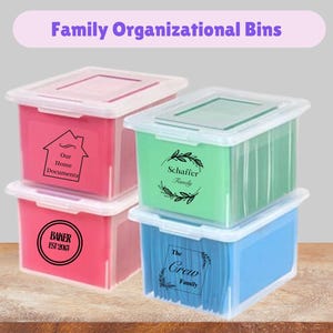 May include: Four rectangular plastic storage bins with clear lids, stacked in two columns. The bins are pink, green, and blue. Each bin features black text and graphics: 'Our Home Documents', 'Schaffer Family', 'BAKER EST 2015', and 'The Crew Family'.