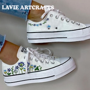 Peut inclure: Baskets en toile blanches avec bordures noires et semelles compensées. Les chaussures sont ornées de broderies florales bleues. Le texte "LAVIE ARTCRAFTS" est visible sur le dessus.