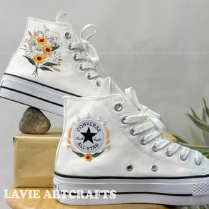 Może przedstawiać: Białe, wysokie trampki Converse z haftowanymi słonecznikami i kłosami pszenicy po bokach. Logo Converse jest haftowane po drugiej stronie z napisem "Chuck Taylor" i "All Star".