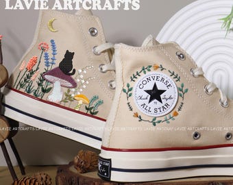 May include: A pair of beige high-top Converse sneakers with embroidered floral designs, a black cat, mushrooms, and a crescent moon. The Converse logo is also embroidered with a floral wreath.