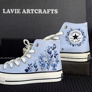 Puede incluir: Zapatillas altas azul claro con diseños bordados de flores y mariposas. Las zapatillas tienen cordones blancos, suela negra y blanca y el logo Converse All Star. El texto "LAVIE ARTCRATS" es visible.