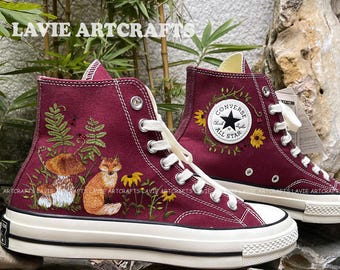 May include: Burgundy high-top sneakers with white laces and soles. The sneakers feature embroidered forest scenes with a fox, mushrooms, and sunflowers. The Converse All Star logo is on the side. The text "LAVIE ARTCRAFTS" is visible.