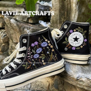 Könnte beinhalten: Ein Paar schwarze Converse-Sneaker mit hohem Schaft und weißen Sohlen. Die Sneaker sind mit gestickten Blumen in den Farben Lila, Blau und Grün verziert. Der linke Sneaker hat das Converse-Logo und den Text "Chuck Taylor All Star" auf der Seite gestickt.