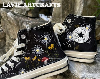 Puede incluir: Zapatillas Converse negras de caña alta con margaritas, flores silvestres y un hongo bordados en el lateral. El logotipo de Converse es visible en el lateral de la zapatilla.