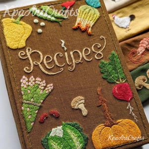 May include: A brown fabric recipe book with embroidered vegetables and the word "Recipes" in cursive. The cover features a yellow bell pepper, green beans, a pumpkin, and other colorful embroidered food items.