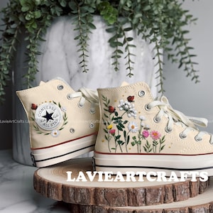 May include: Cream high-top trainers with embroidered floral and insect designs. The trainers feature a Converse logo patch and are displayed on a wooden slice. The embroidery includes daisies, ladybirds, and bees.