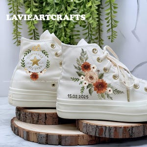 May include: Cream-colored high-top sneakers with embroidered floral designs in shades of orange, brown, and green. The left shoe features the Converse All Star logo and a crown. The right shoe has the date 15.02.2025. Gold eyelets and laces complete the look.