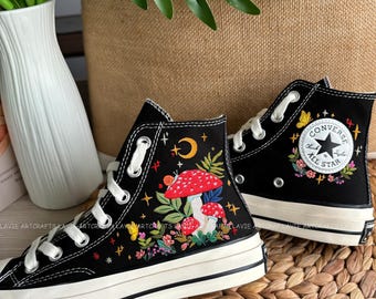 May include: Black high-top Converse sneakers with a whimsical embroidered design featuring a mushroom, a snail, a crescent moon, and stars. The design is surrounded by colorful flowers and leaves. The Converse logo is visible on the side of the shoe.