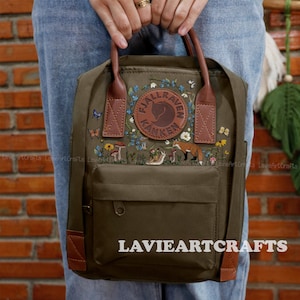 May include: Olive green backpack with brown leather accents and embroidered forest scene. Features a front pocket and top handle. The backpack has the text "FJALLRAVEN KANKEN" and "LAVIEARTCRAFTS".