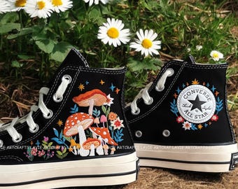May include: Black high-top Converse sneakers with embroidered mushrooms, flowers, and stars. The Converse logo is also embroidered with colorful flowers. The shoes are on a brown surface with white daisies.