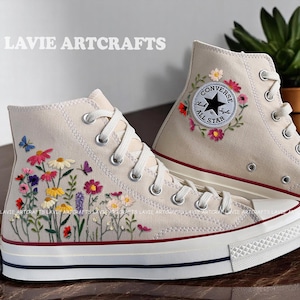 Puede incluir: Zapatillas altas color crema con diseños florales y mariposas bordadas. La zapatilla izquierda presenta un jardín de flores colorido, la derecha tiene un parche Converse All Star rodeado de flores. El texto "LAVIE ARTCRAFTS" está impreso en el lateral.