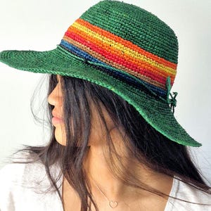 Pode incluir: Um chapéu de sol de crochê verde com uma faixa de arco-íris. O chapéu tem uma aba larga e é feito de um material natural.