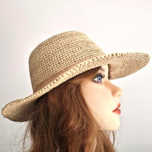 Könnte beinhalten: Beigefarbener Strohhut mit breiter Krempe und beigem Band. Der Hut hat eine klassische Fedora-Form und ist für den Sonnenschutz konzipiert. Der Hut ist neutralfarben und kann zu vielen Outfits getragen werden.