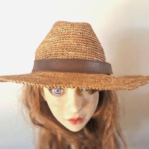 Pode incluir: Um chapéu de palha tecido com uma faixa de couro castanho. O chapéu tem uma aba larga e uma forma de fedora clássica. O chapéu é de cor bege claro e parece ser feito à mão.