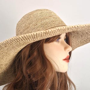 Puede incluir: Un sombrero de sol de ala ancha hecho de paja tejida. El sombrero tiene una copa redondeada y un ala ancha y suavemente inclinada. El color beige natural de la paja le da al sombrero un aspecto veraniego e informal. El sombrero está diseñado para dar sombra.