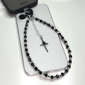 May include: A black and silver phone charm with a cross pendant and black beads. The charm is attached to a white smartphone.