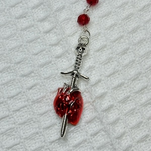 May include: A necklace featuring a silver sword piercing a red heart pendant. The sword has a skull at the top. The necklace also includes red beads. The background is a white, textured surface.