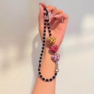 May include: A beaded phone charm featuring three heart-shaped beads with animal print designs in yellow, pink, and purple. The charm has black beads and a silver-tone clasp.