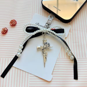 May include: A phone charm featuring a silver-toned metal cross pendant with a rose design, attached to a black satin ribbon bow. The bow is accented with white faux pearl beads and a silver lobster clasp, mounted on a white display card.