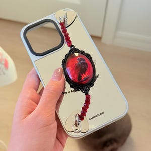 May include: A reflective silver phone case featuring a black camera cutout and the text CASENOTCAKE. Attached is a decorative charm with two silver heart-shaped anchors, red crystal beads, and a central black oval frame containing a red bird graphic.