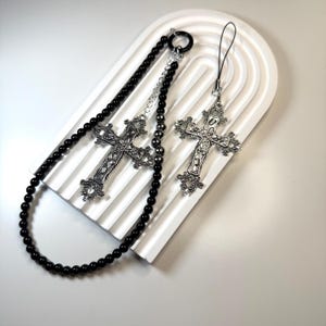 May include: A necklace and a charm featuring ornate silver crosses. The necklace has a strand of black beads. The charm has a black cord. The crosses have detailed floral and scrollwork designs. The items are displayed on a white, textured surface.