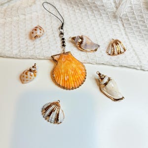May include: An assortment of seashell charms and pendants. The central pendant is a large, orange scallop shell with gold trim, suspended from a black cord. Other shells include white, gold, and speckled designs. The items are arranged on a white surface.