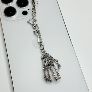 May include: A silver skeleton hand charm attached to a silver chain. The charm is dangling from a phone case.