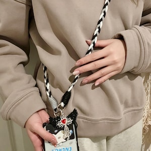 May include: A black and white braided phone strap with a decorative phone case. The phone case has a black and white design with a cross and wings charm. The strap is attached to the phone case with a silver ring.