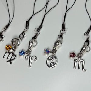 May include: Close-up of several silver-toned zodiac charm phone straps. Each charm features a different zodiac symbol, such as Virgo, Aries, Cancer, and Libra, with a colorful gemstone accent. The straps are black.