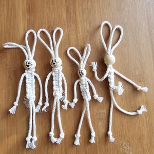 Peut inclure: Quatre décorations de lapin en ficelle blanche avec des têtes en bois. Les lapins ont des yeux noirs et un sourire noir. Les lapins sont de tailles différentes et ont des poses différentes.