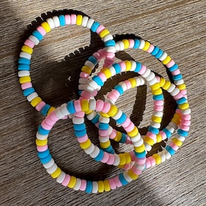 Könnte beinhalten: Ein Set aus drei kreisförmigen Armbändern, bestehend aus kleinen, glatten, zylindrischen Perlen in einem sich wiederholenden Muster aus Weiß, Hellrosa, leuchtendem Gelb und Himmelblau. Die Perlen sind auf ein elastisches Band gefädelt.