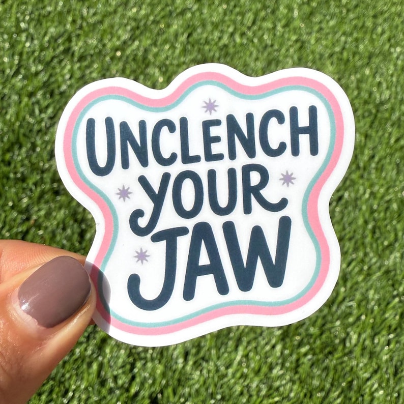 K&ouml;nnte beinhalten: Ein wei&szlig;er Aufkleber mit den Worten "UNCLENCH YOUR JAW" in Dunkelblau, umgeben von einem rosa und hellblauen Rand. Der Aufkleber hat eine wolkenartige Form und kleine Sternakzente.