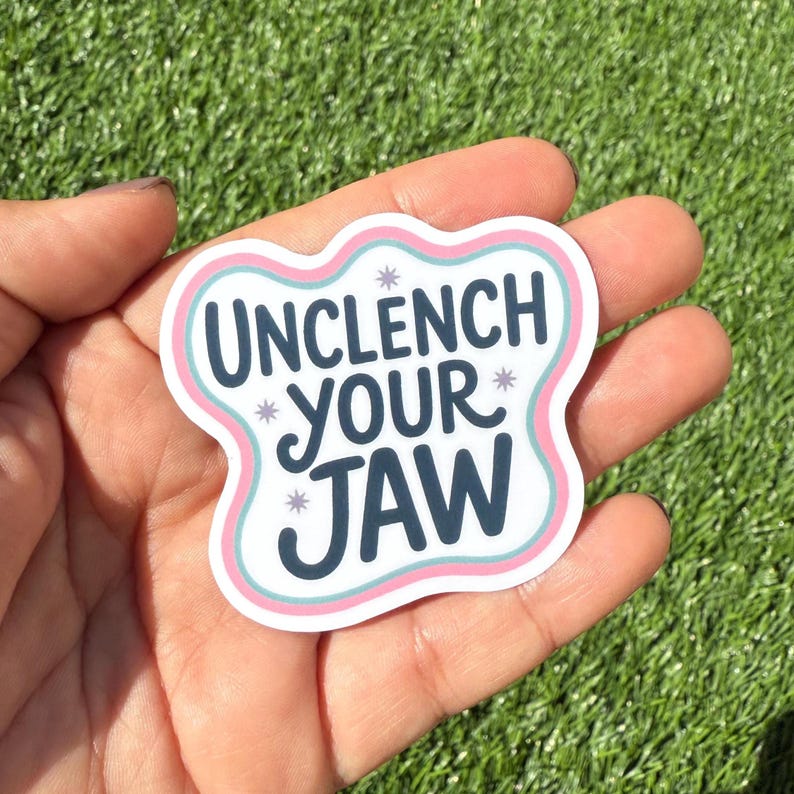 K&ouml;nnte beinhalten: Ein wei&szlig;er Aufkleber mit dem Text "UNCLENCH YOUR JAW" in Dunkelblau, umrandet von hellblau und rosa. Kleine Sternakzente sind um den Text herum verstreut. Der Aufkleber wird in einer Hand vor einem gr&uuml;nen Hintergrund gehalten.