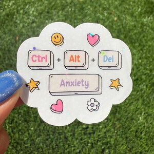 May include: A white sticker with a cloud shape and the text "Ctrl + Alt + Del = Anxiety." The sticker has a smiley face, a pink heart, a star, and a flower. The sticker is held by a person with blue nail polish.