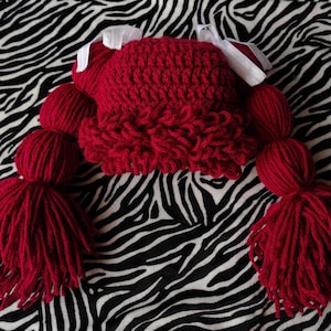 Puede incluir: Un gorro rojo tejido a mano con ganchillo con coletas de hilo adjuntas y lazos blancos. El gorro presenta una puntada texturizada y está diseñado para parecerse al peinado de un personaje. Las coletas están hechas de hilo grueso.