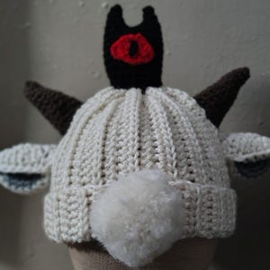 May include: A cream-colored crochet hat with a black and red eye design on top, brown horns, and white ears. A fluffy white pom-pom is attached to the back of the hat. The hat is designed to resemble a goat.