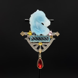 May include: An elaborate brooch with a light blue horse head carving. It features a rectangular aquamarine stone, diamonds, and colourful gemstones. A teardrop-shaped red gemstone hangs from the bottom. The brooch is set against a black background.