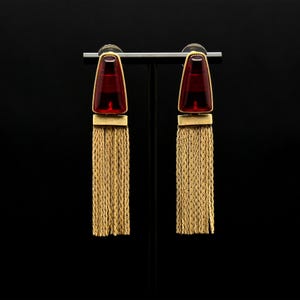 Puede incluir: Un par de pendientes de oro con piedras de vidrio rojo en forma de lágrima y largos flecos de cadena dorada.