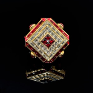 Może przedstawiać: Pierścionek w kształcie kwadratu z czerwonym kamieniem szlachetnym otoczonym mniejszymi, przezroczystymi kamieniami szlachetnymi. Pierścionek jest osadzony w złocie i ma geometryczny wzór.