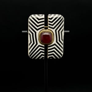 May include: A geometric brooch with a black and white zigzag pattern. The brooch has a rectangular shape with a central red gemstone set in a gold-coloured frame. The design is bold and modern.