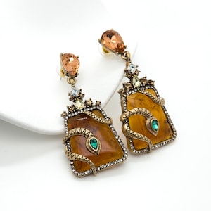 Puede incluir: Un par de pendientes ornamentados con un colgante rectangular de color ámbar con un diseño de serpiente dorada y una gema melocotón en forma de lágrima. Los pendientes tienen una estética vintage, con pequeños cristales transparentes y un acento de gema verde.