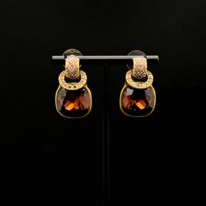 May include: A pair of gold-toned earrings with large, square-cut, brown-colored gemstones. The earrings have a smaller, circular accent above the main stone, embellished with smaller, pink-toned stones. The earrings are displayed on a black stand.