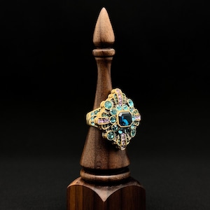 Op de afbeelding: Een gouden ring met een grote blauwe edelsteen omringd door kleinere blauwe, groene en paarse edelstenen.