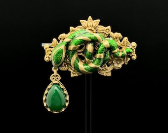 Puede incluir: Broche dorado con un diseño de serpiente esmaltada en verde y oro. La serpiente se enrolla alrededor de un motivo floral, con un colgante de piedra verde en forma de lágrima. El fondo es negro.